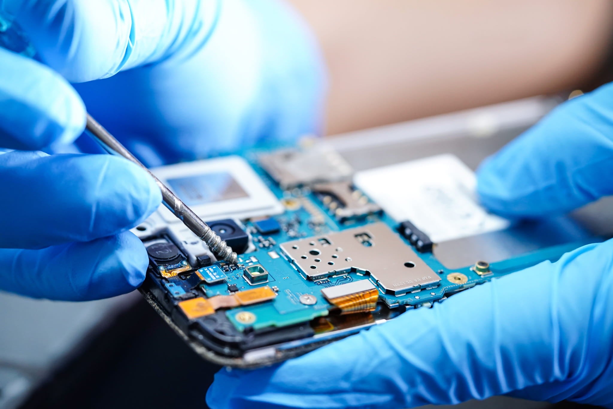 Person wearing blue gloves working on a smartphone circuit board.